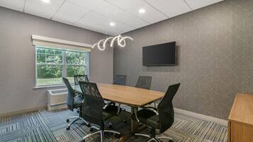 A conference room with a large table, four chairs, a flat-screen TV, and a window with a view of greenery.