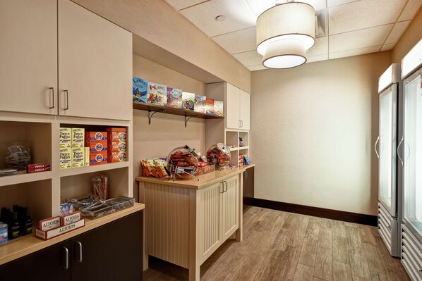 A hotel pantry with shelves stocked with snacks and beverages, a counter with food items, and a view of the hallway.