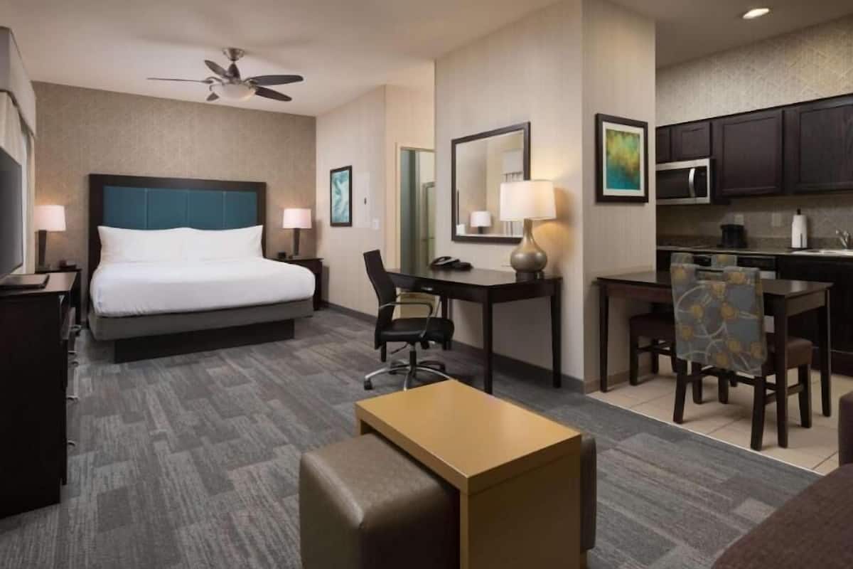 A hotel room with a bed, desk, chair, kitchenette, and a ceiling fan.