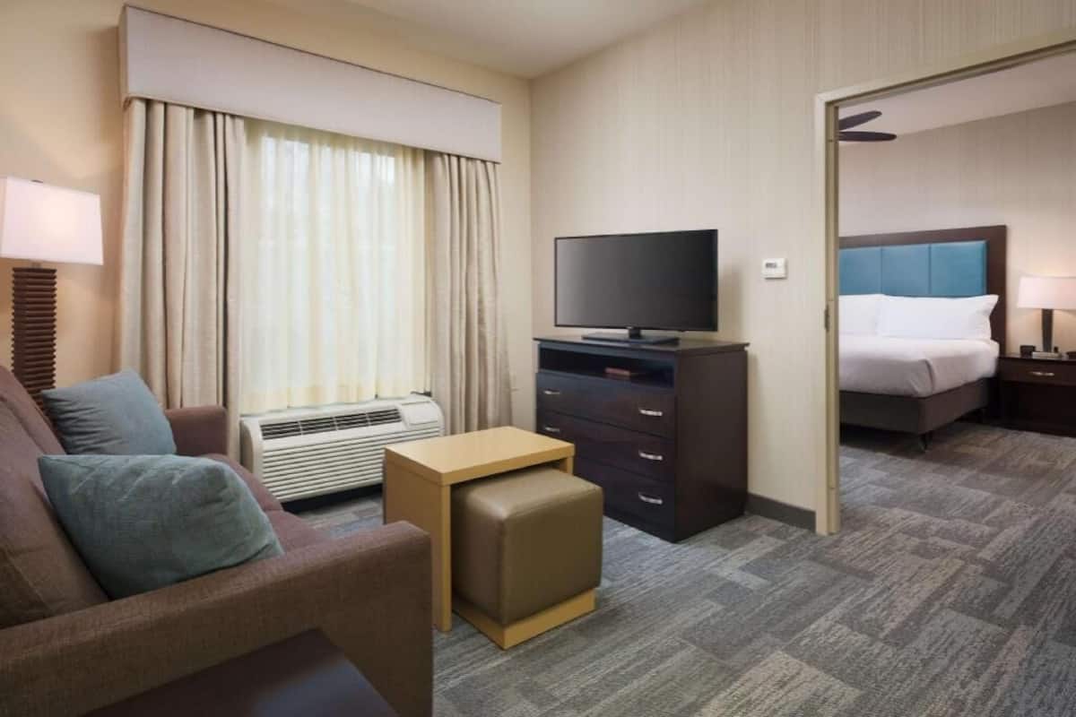 A hotel room with a sofa, a television on a stand, a small table, an air conditioning unit, and a bed with a headboard.