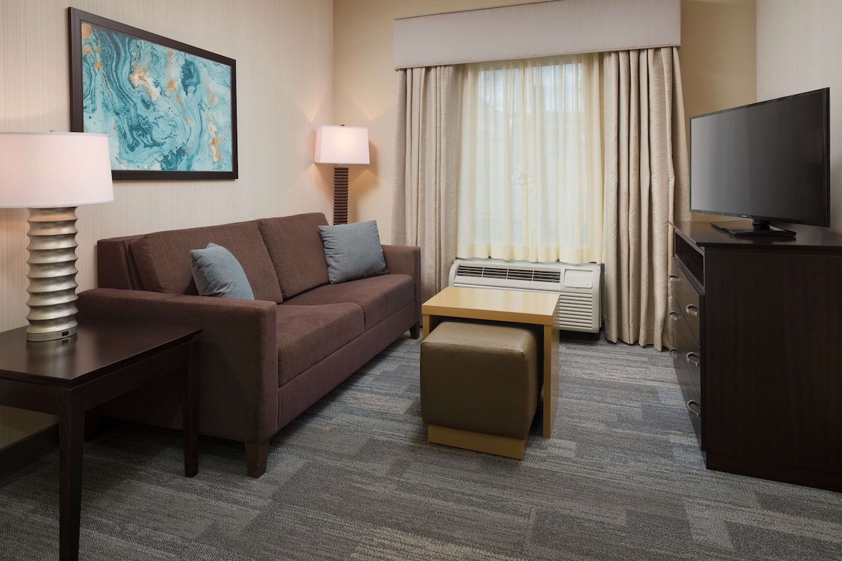 A hotel room with a brown sofa, a small table, a TV, a lamp, and a painting on the wall.