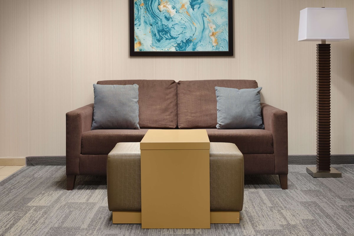 A modern living room with a brown sofa, two grey cushions, a small beige ottoman, and a framed abstract painting on the wall.