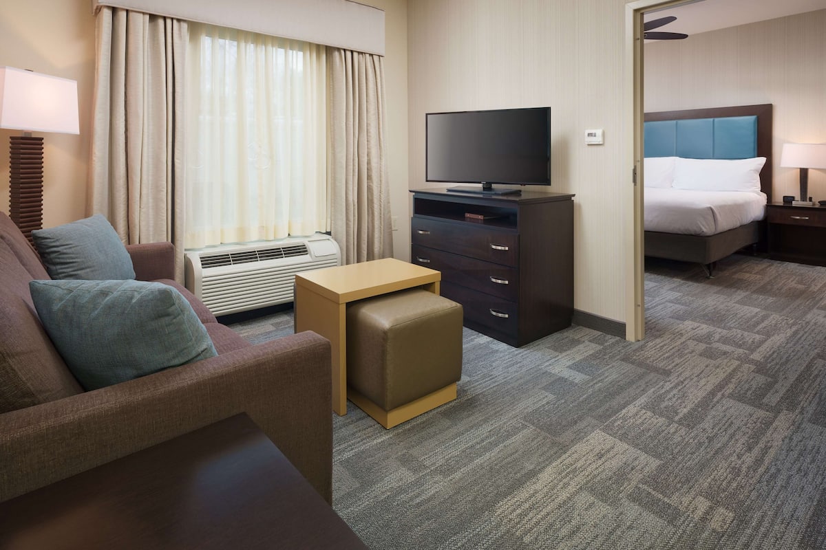 A hotel room with a sofa, a TV on a stand, a small table, an air conditioning unit, and a bed with a headboard.