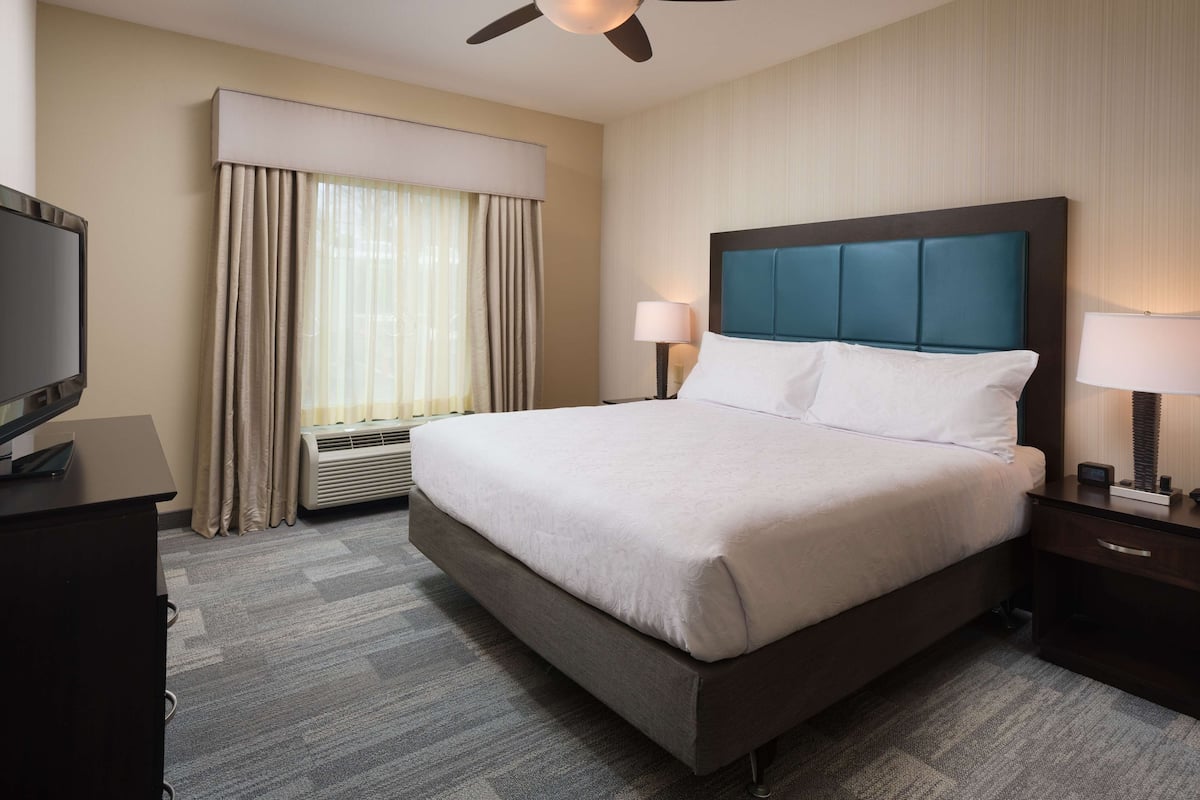 A hotel room with a large bed, a flat-screen TV, a ceiling fan, a window with curtains, and a nightstand with a lamp.