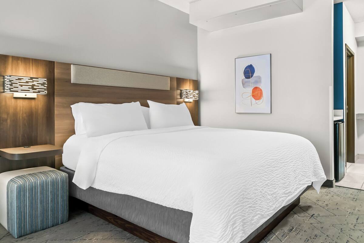 A modern hotel room with a large bed, a bedside table, a wall-mounted lamp, and a framed abstract painting.
