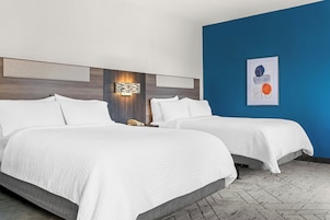 Habitación de hotel con dos camas, una mesita de noche, una lámpara fijada a la pared y un cuadro abstracto enmarcado.