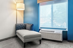 Een hoekkamer met een grijze, beklede zitbank, een wandlamp en een raam met uitzicht op bomen.
