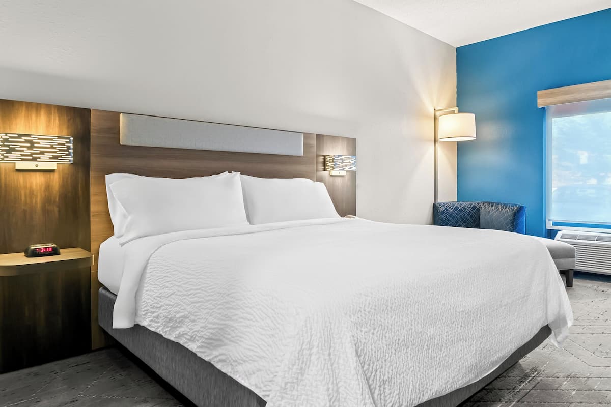 A modern hotel room with a large bed, a nightstand with a clock, a wall-mounted light, and a window with a view.