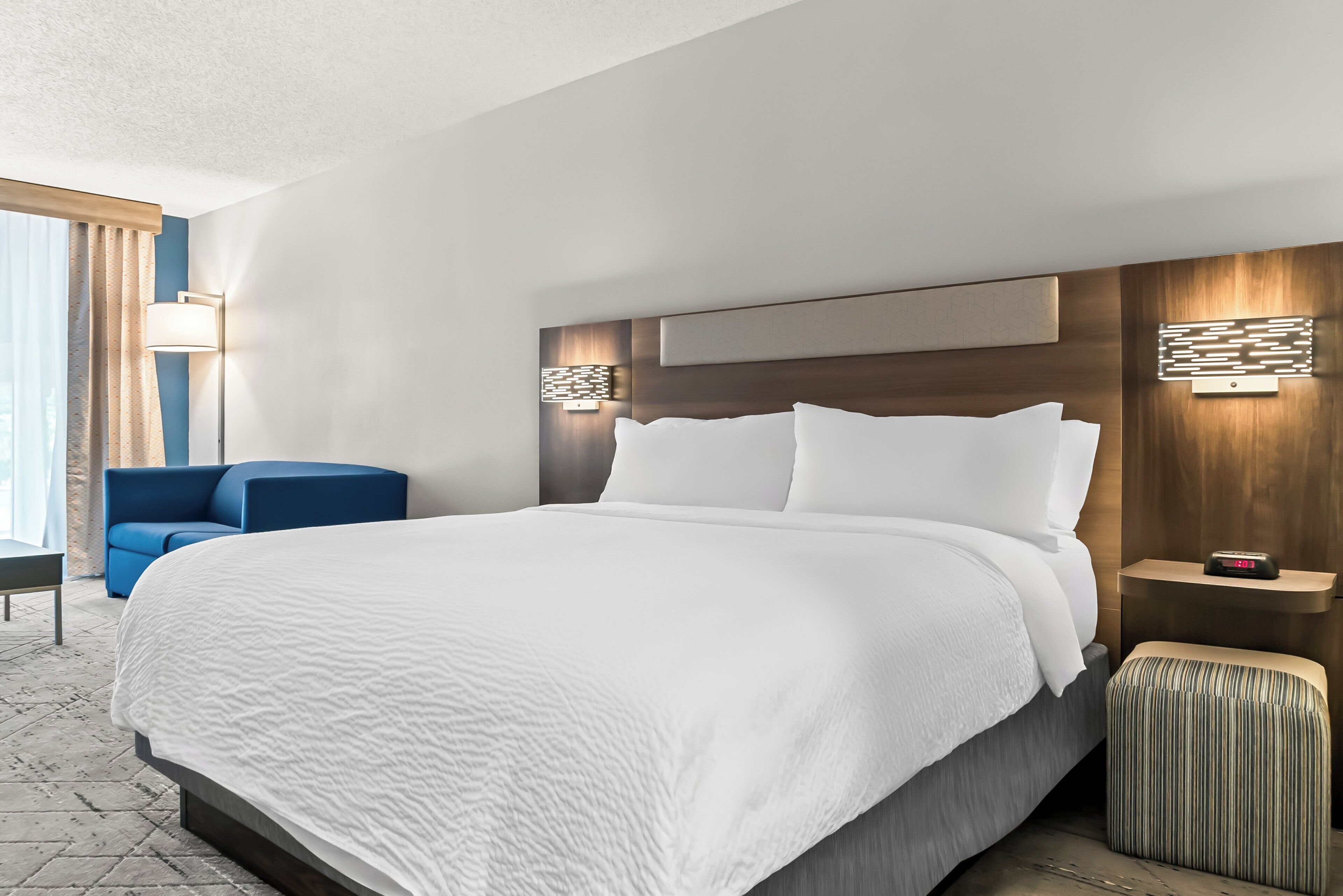 Een moderne hotelkamer met een groot bed, een blauwe fauteuil, een nachtkastje met een klok en een wandlamp.