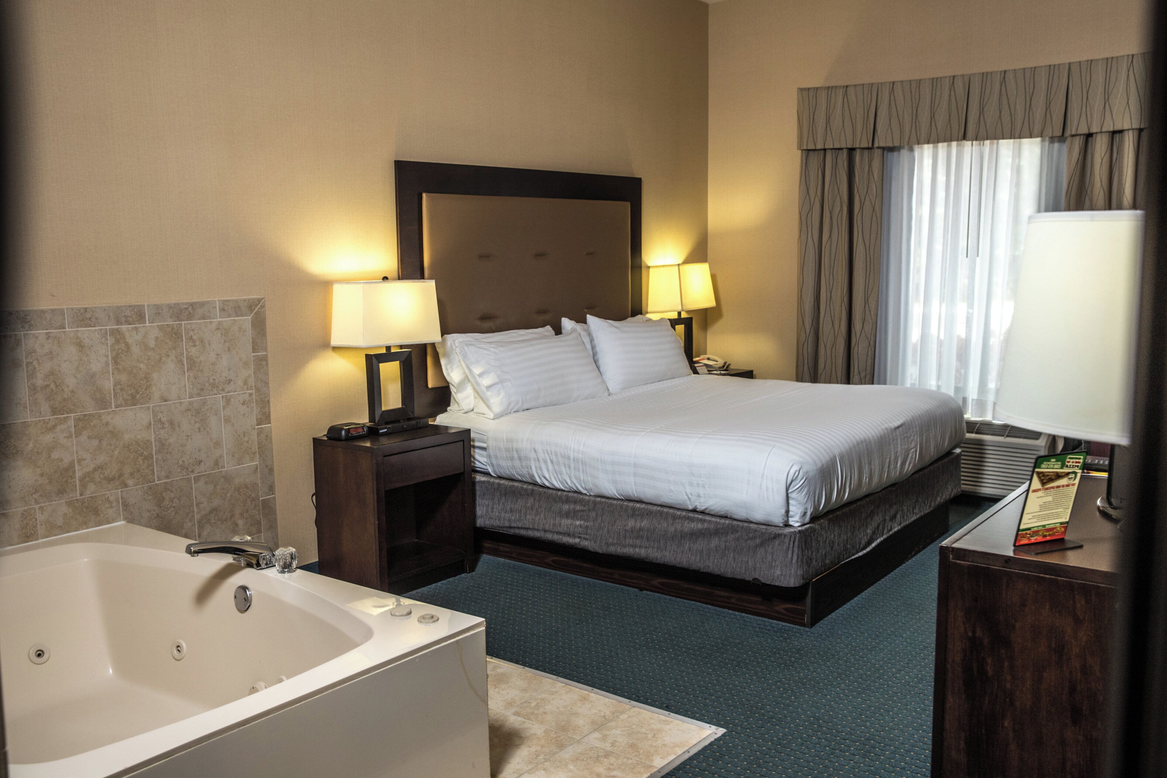 Quarto de hotel com uma cama grande, duas luminárias de cabeceira, uma mesa de cabeceira, um banheiro com banheira e uma janela com cortinas.