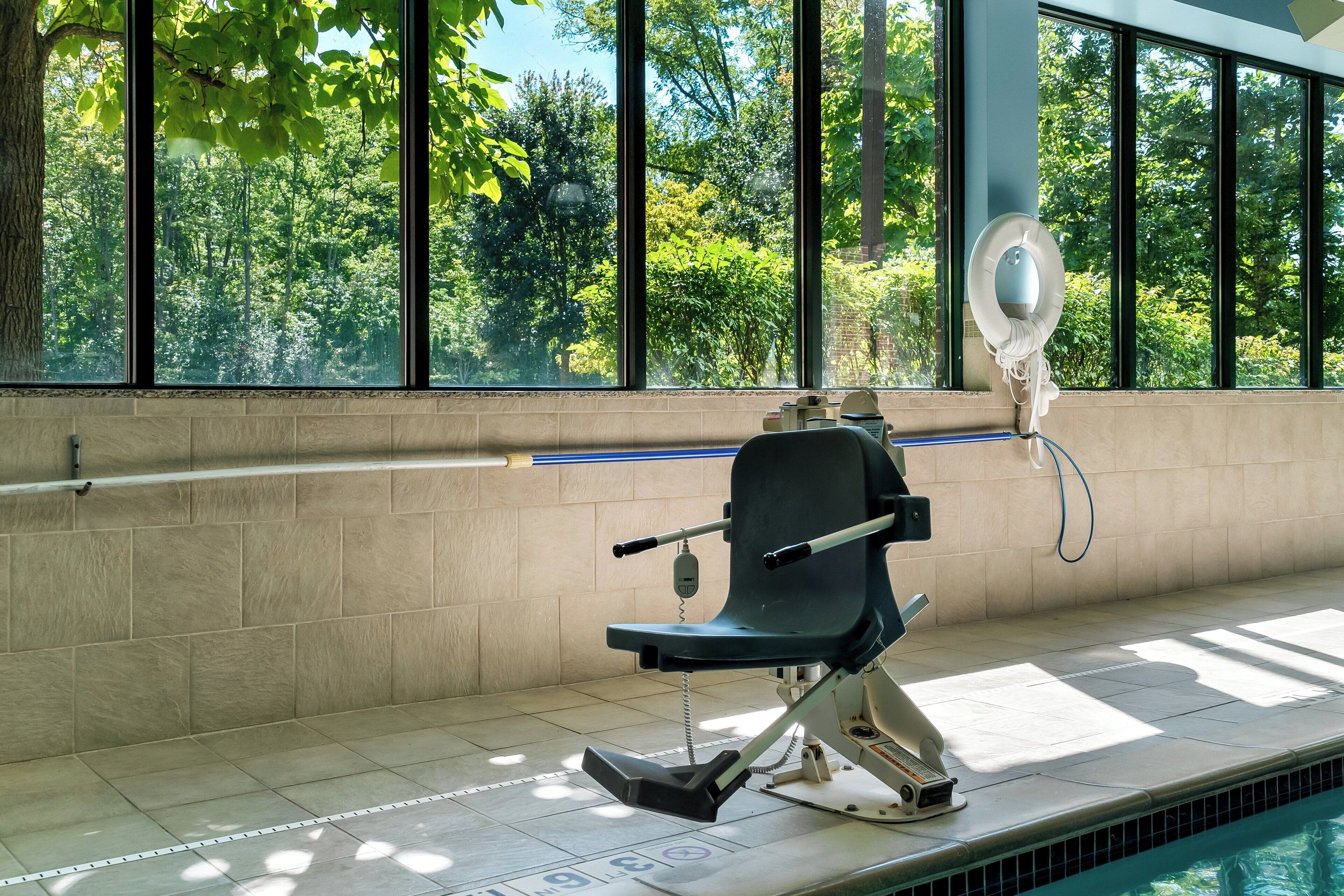 A poolside area with a wheelchair-accessible bench, a fan, and a view of trees through large windows.