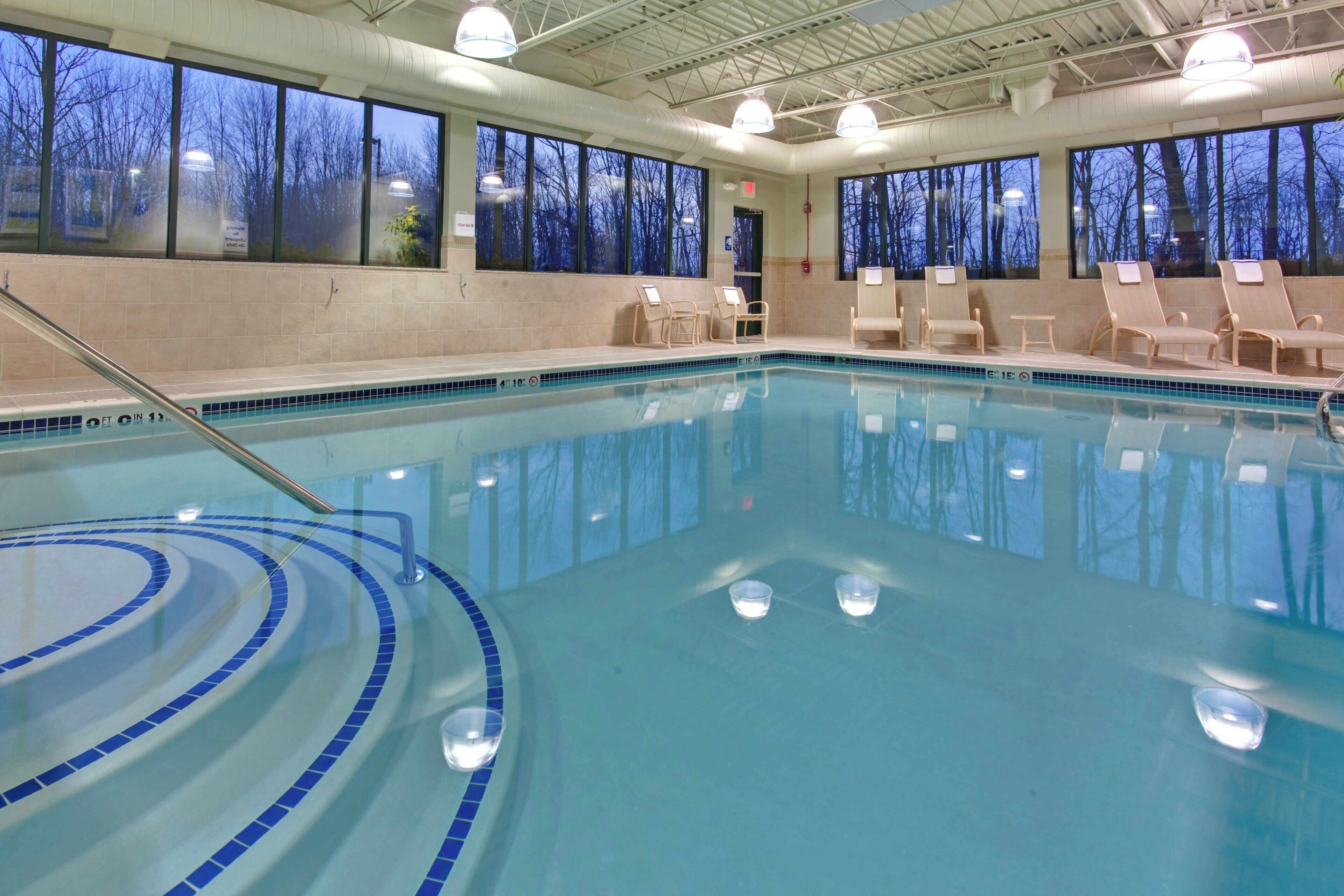 An indoor swimming pool with clear blue water, surrounded by lounge chairs and large windows.