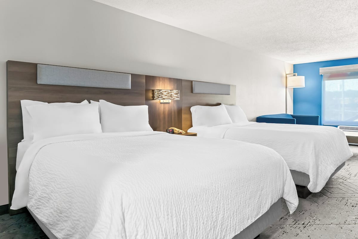 A hotel room with two beds, a modern headboard, a bedside table with a lamp, and a large window with a view.