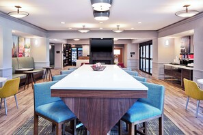 A modern hotel dining area with a long table, blue upholstered chairs, a central fireplace, and a bar area. - Hampton Inn Detroit/Utica-Shelby Township (Utica)