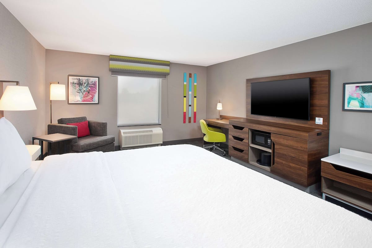 A hotel room with a large bed, a desk with a flat-screen TV, a chair, a small table, and a window with blinds.