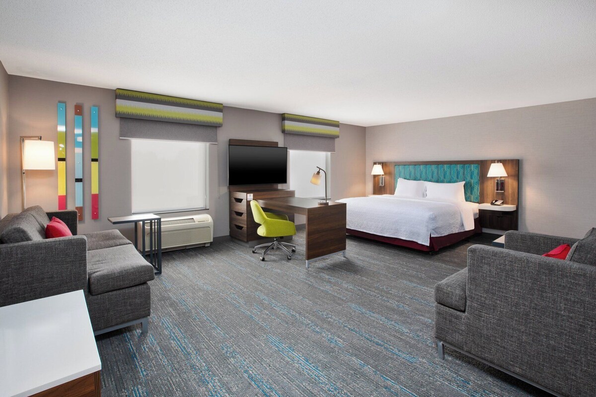 A hotel room with a bed, two armchairs, a desk, a TV, and a window.