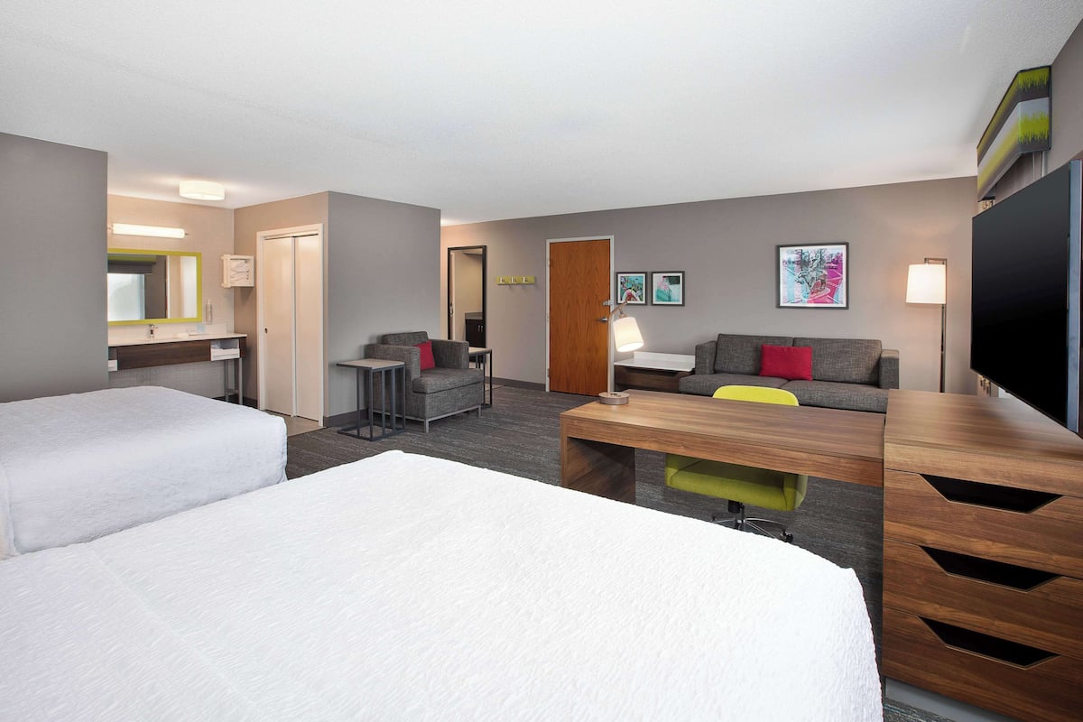 A hotel room with two beds, a desk, a sofa, a TV, and a bathroom with a mirror and sink.