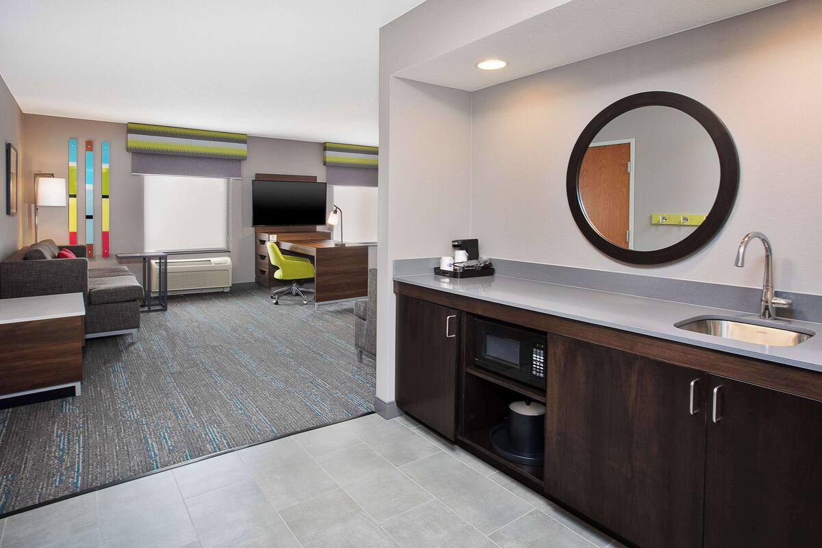 A modern hotel room with a kitchenette, a seating area with a sofa and armchairs, a flat-screen TV, and a large mirror above the sink.