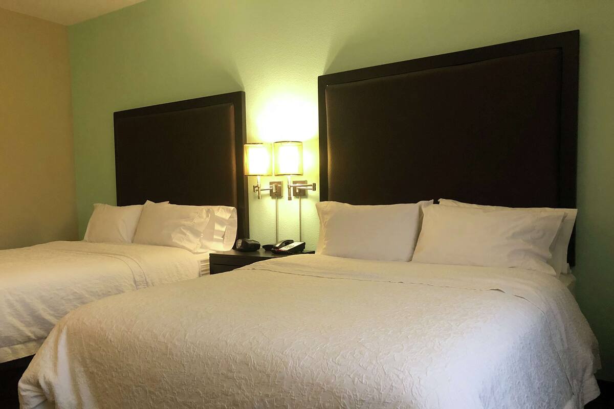 A hotel room with two beds, each with white linens and dark headboards. There is a nightstand with a phone and a lamp between the beds.