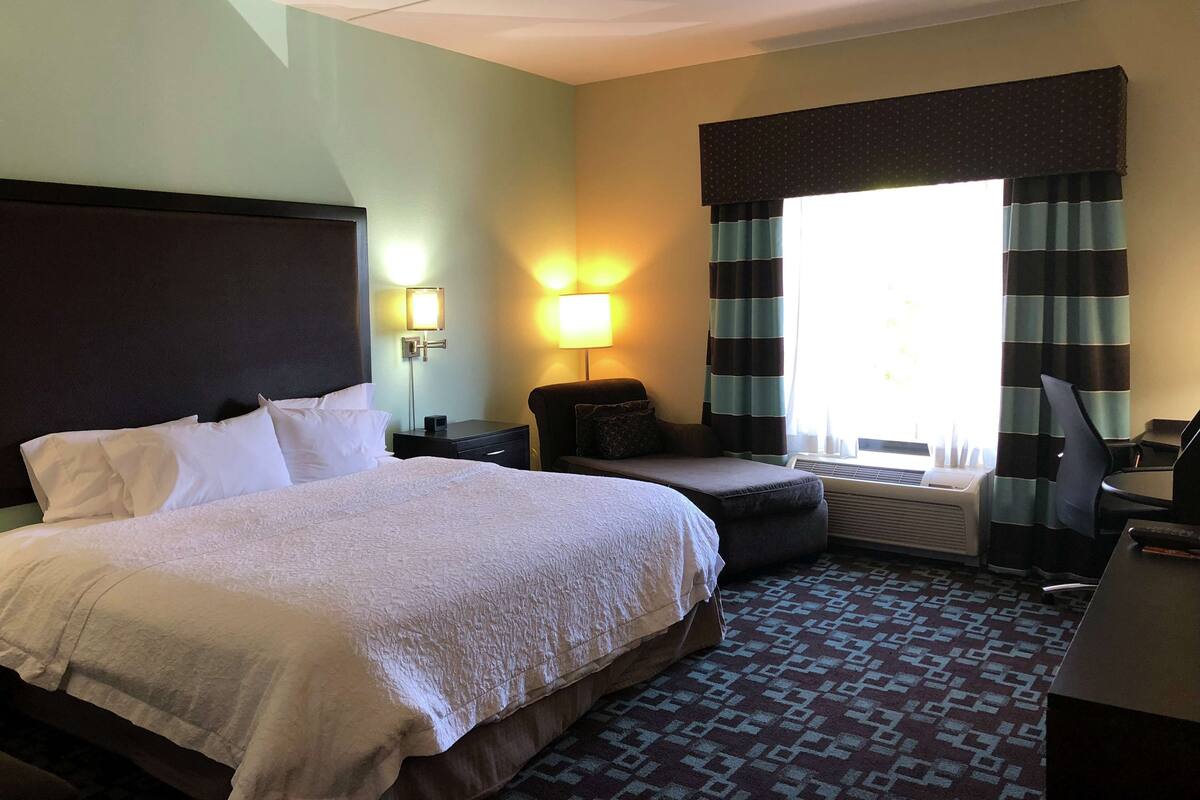 A hotel room with a large bed, a desk, a chair, a lamp, and a window with curtains.