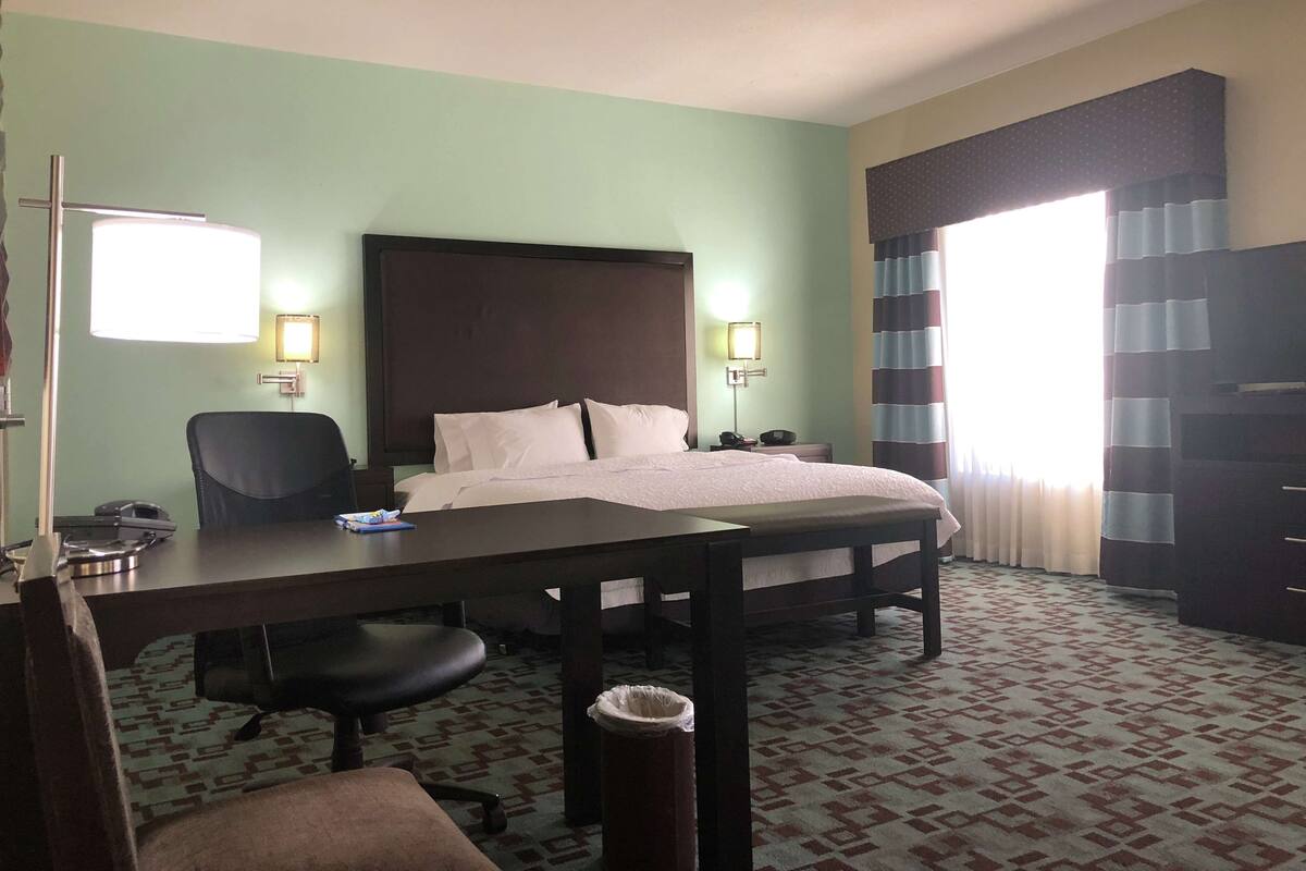 A hotel room with a bed, desk, chair, and a trash bin.