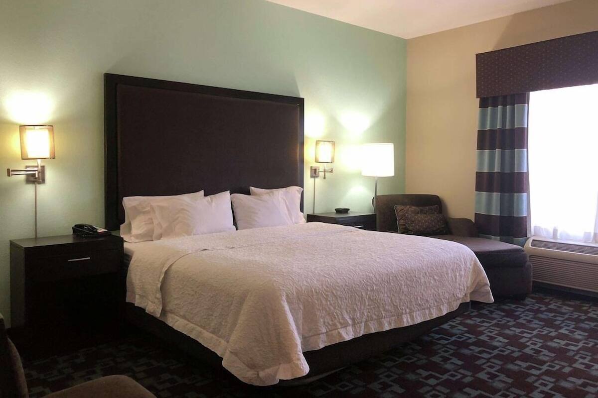 A hotel room with a large bed, bedside lamps, a chair, and a window with curtains.