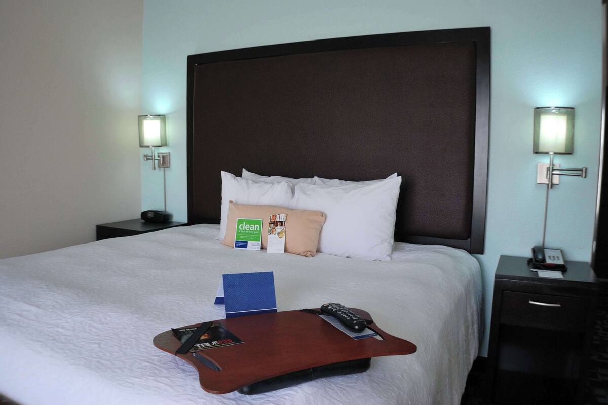 A hotel room with a large bed, two bedside tables, a nightstand, a remote control, a magazine, and a pillow with a 'clean' sign.