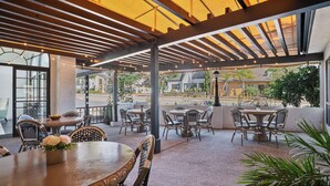 An outdoor patio area with tables and chairs, a pergola structure, and a view of the street and buildings in the background.
