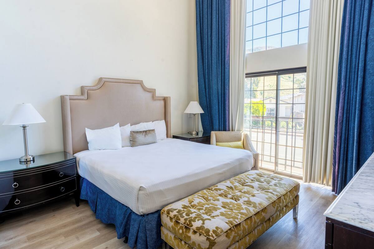 A neatly arranged hotel room with a large bed, bedside tables with lamps, a bench, and a view through a window.