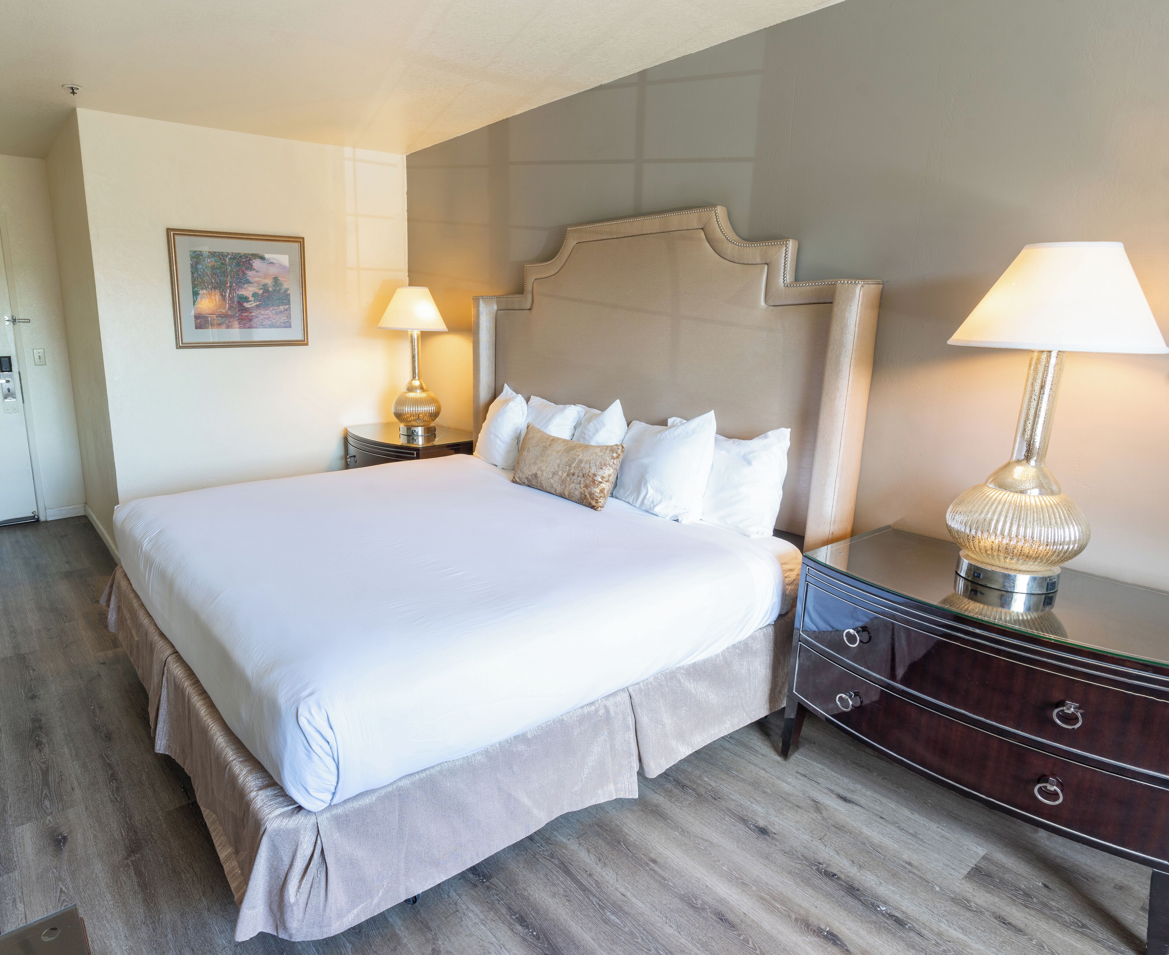 Una cama bien hecha con cabecera, dos mesitas de noche con lámparas y suelo de madera.