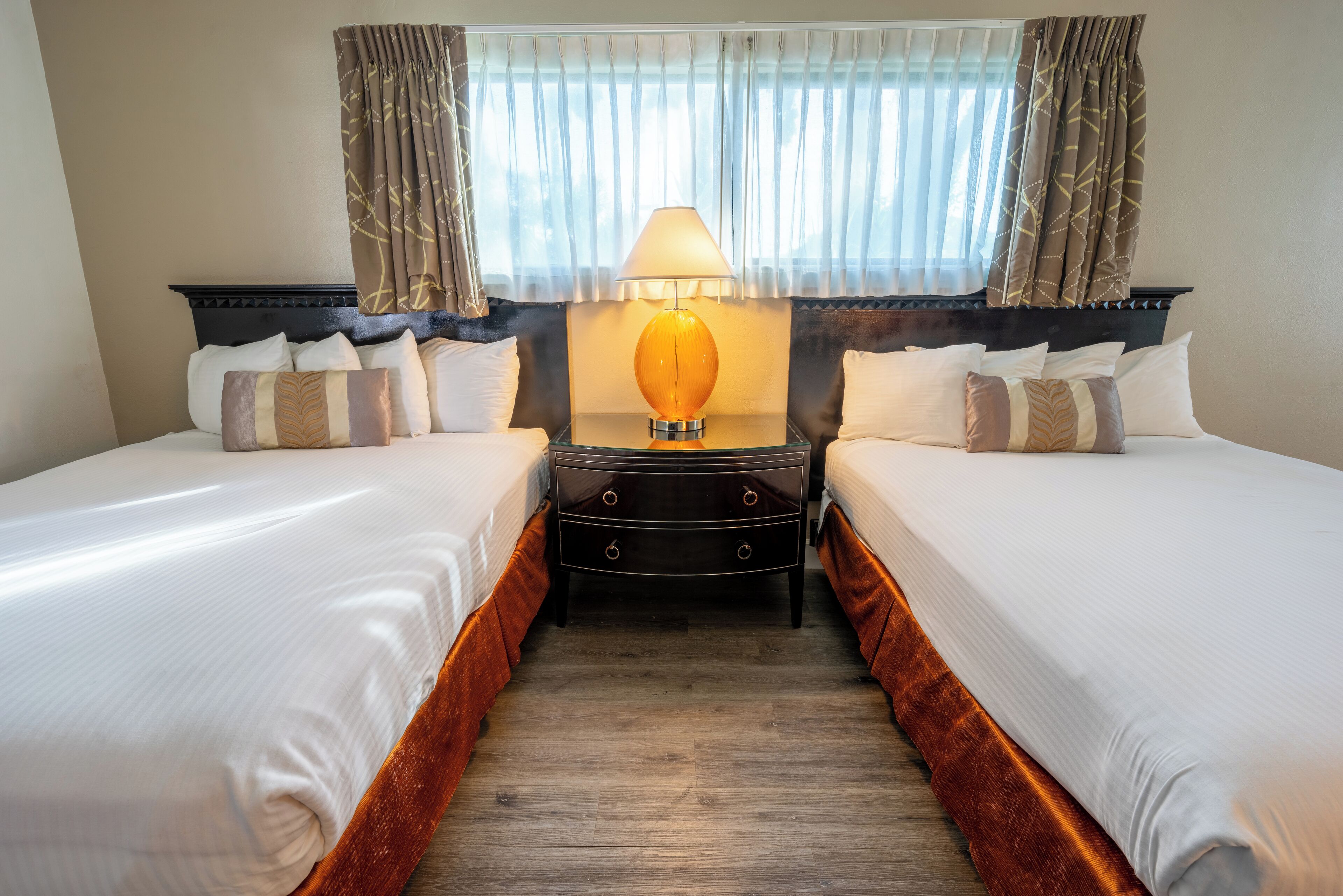 Een hotelkamer met twee bedden, een nachtkastje met een lamp en een raam met gordijnen.