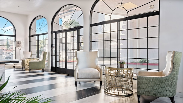 A spacious lobby with large arched windows, a black and white tiled floor, and a mix of modern and classic furniture including a white armchair, a green patterned armchair, and a gold-rimmed round table.