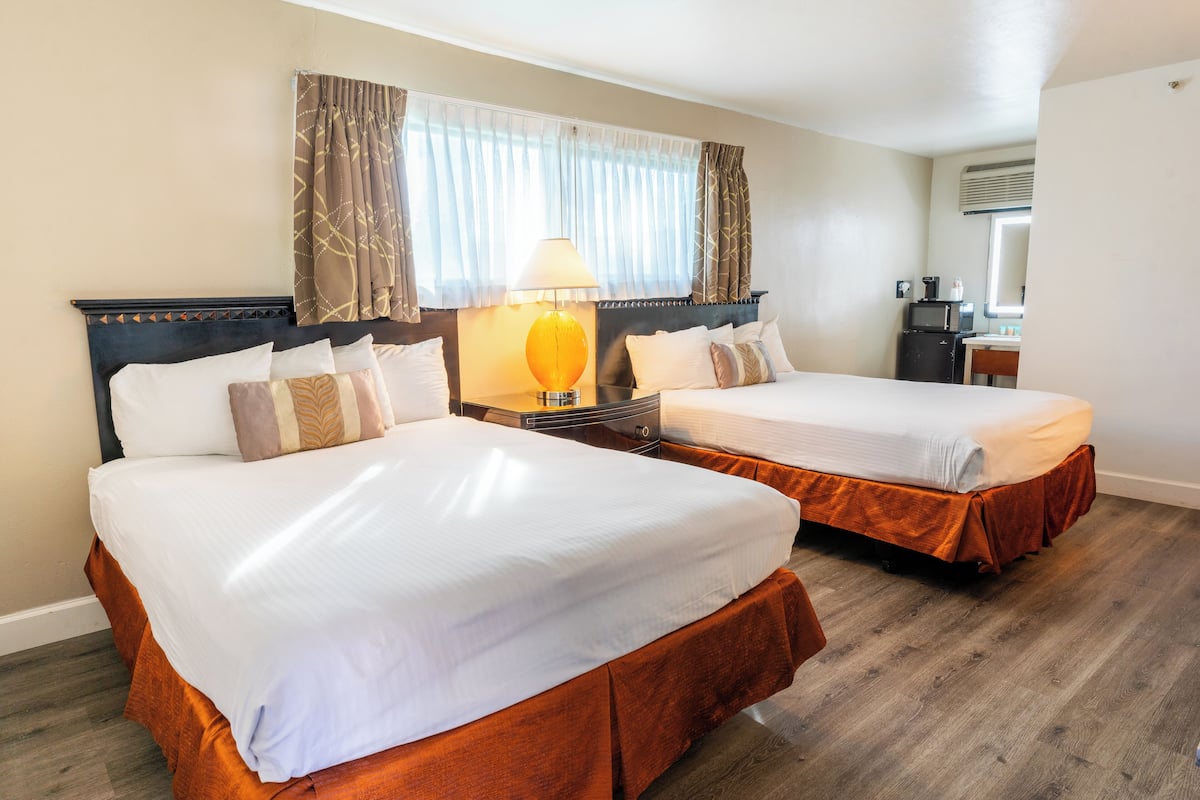 A hotel room with two beds, a nightstand with a lamp, a desk with a computer, and a window with curtains.