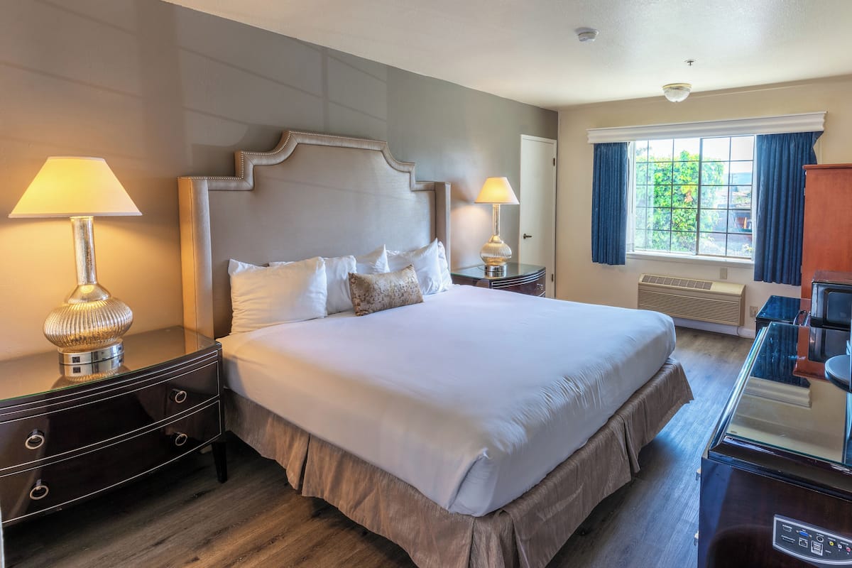 A neatly made bed with white linens, two bedside lamps, a wooden headboard, a nightstand with drawers, a window with curtains, and a built-in heater.