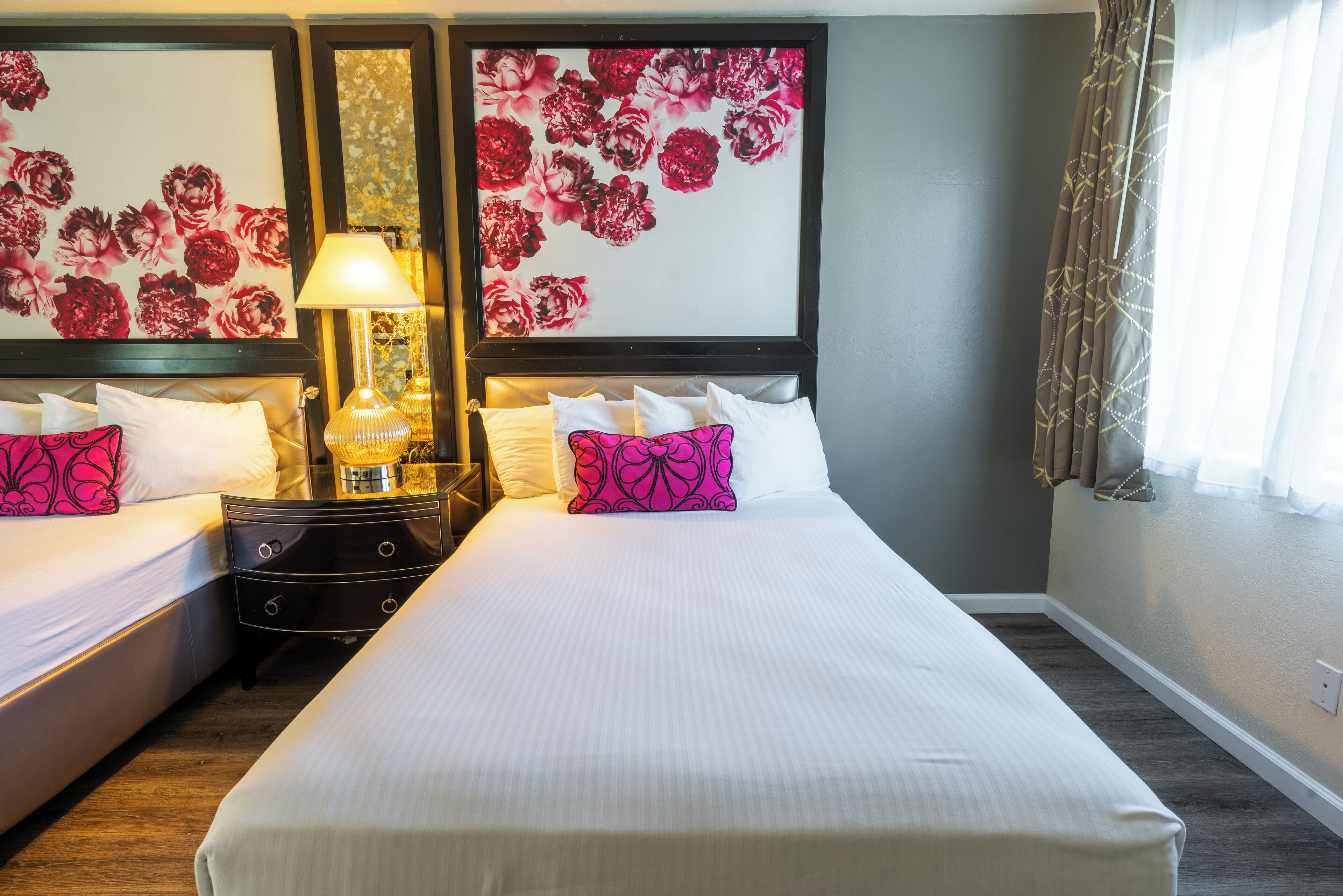 Habitación de hotel con dos camas, una mesita de noche con lámpara y cuadros con motivos florales en las paredes.
