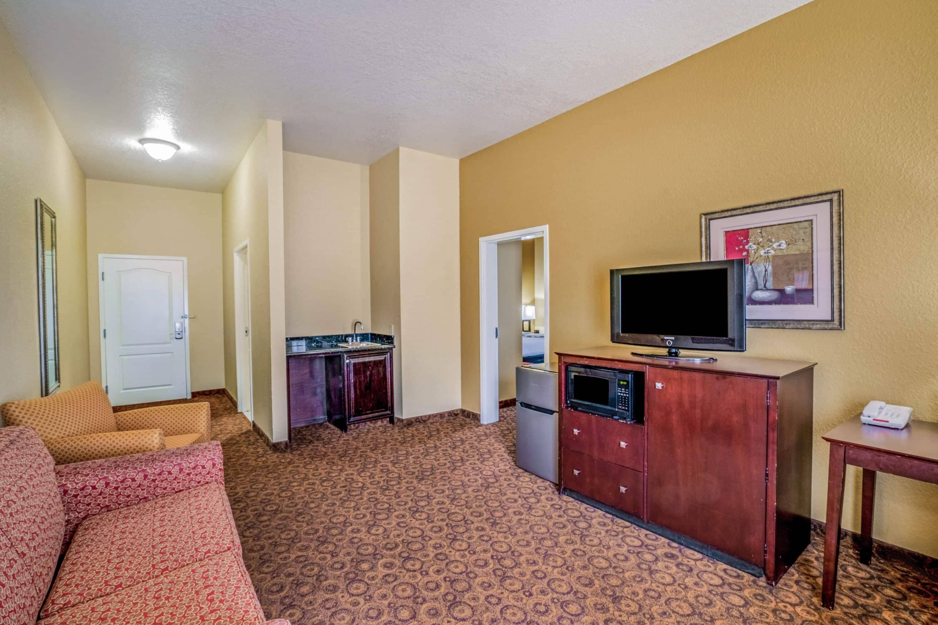 A hotel room with a sofa, a television on a wooden cabinet, a kitchen area with a refrigerator, and a door leading to another room.
