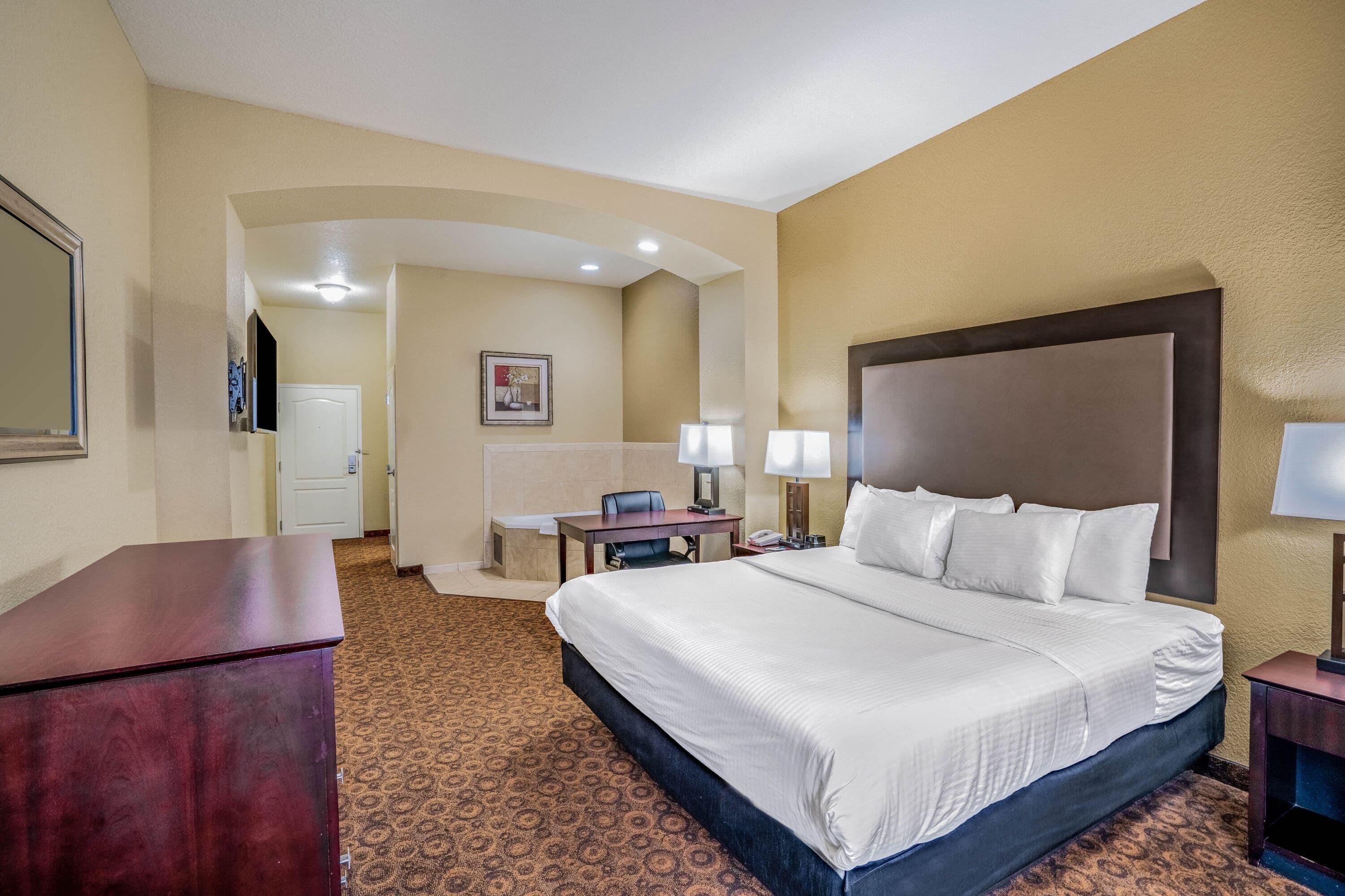 A hotel room with a large bed, a desk, a chair, and a small table with lamps.