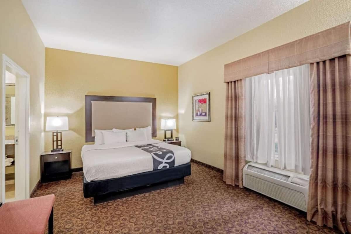 A hotel room with a bed, two bedside lamps, a window with curtains, a wall-mounted air conditioner, and a framed picture on the wall.