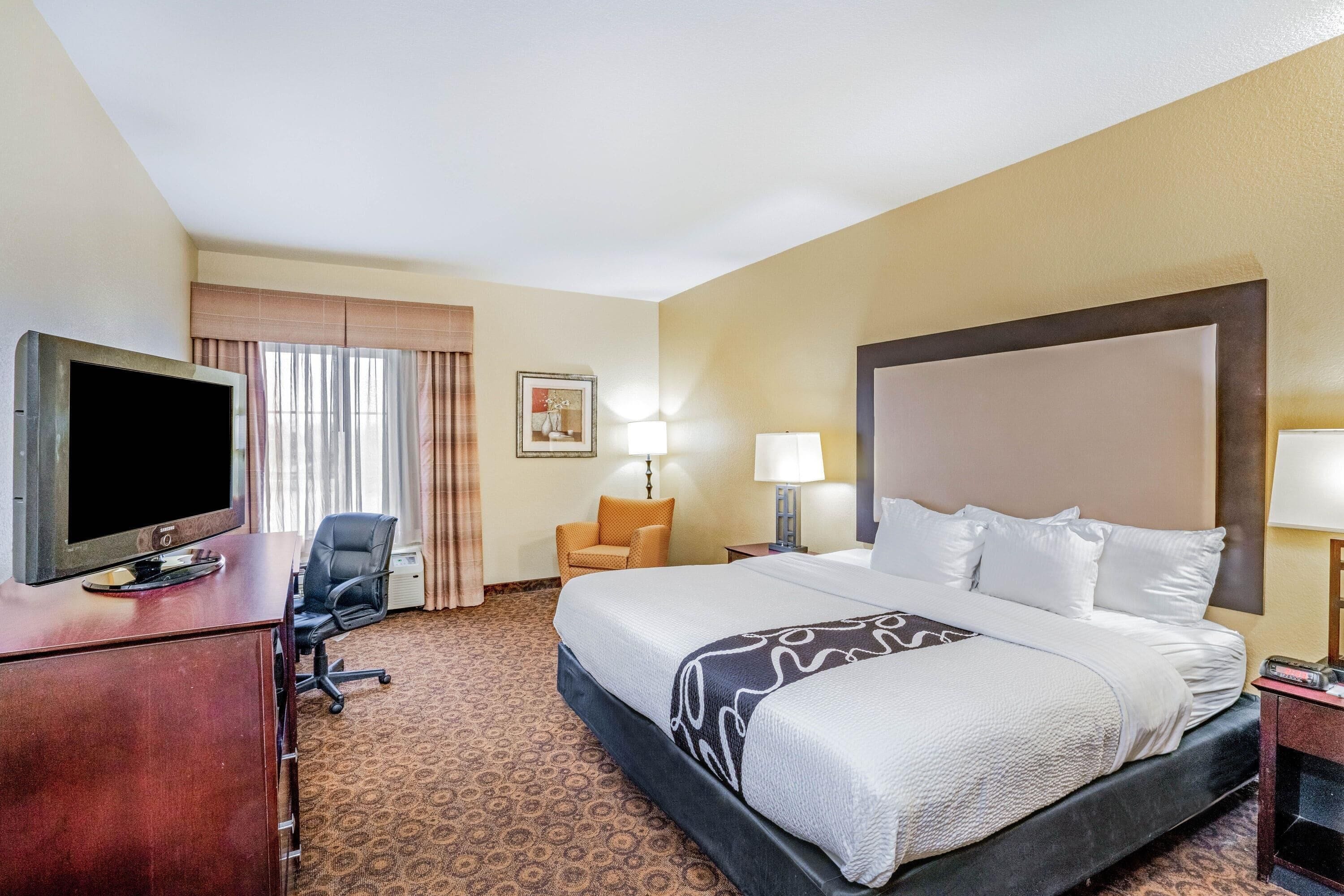 A hotel room with a bed, a desk with a television, a chair, a lamp, and a nightstand.