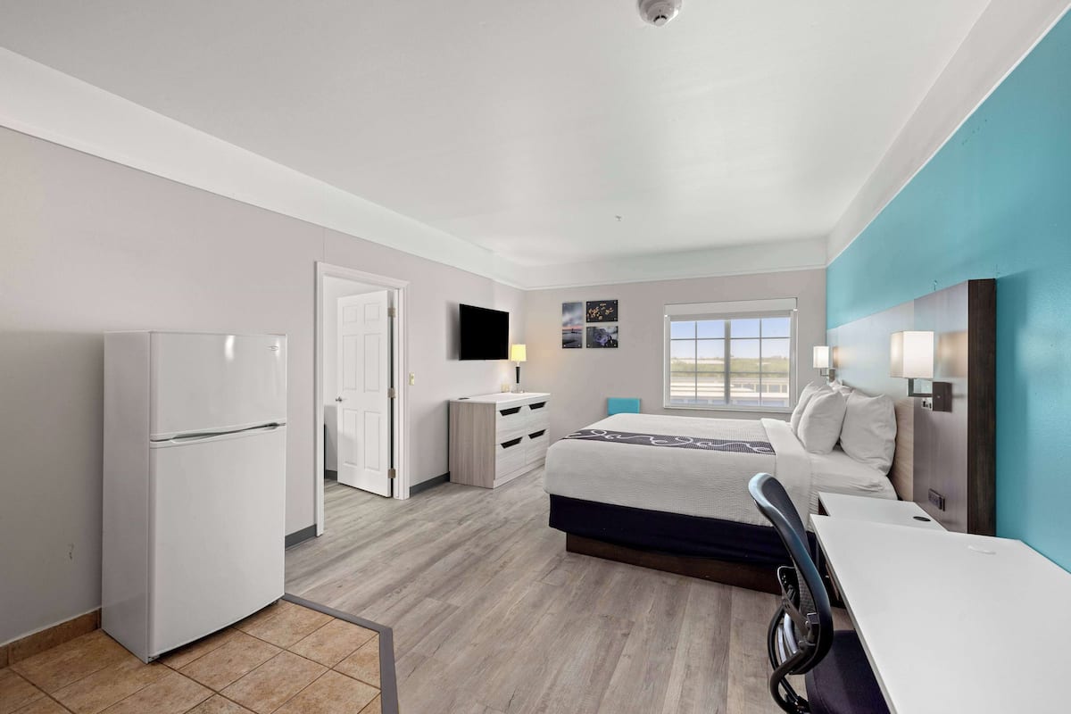 A modern hotel room with a bed, a desk, a refrigerator, a dresser, and a TV.