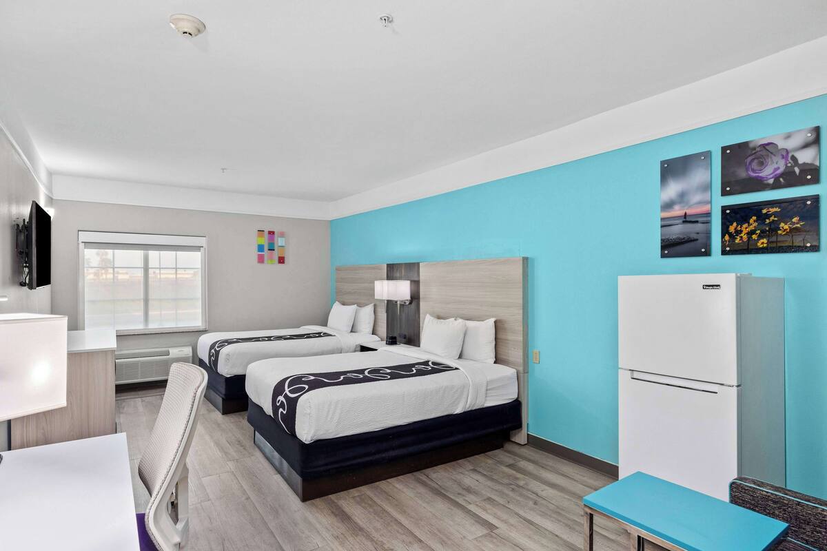 A hotel room with two beds, a desk, a chair, a refrigerator, and artwork on the walls.