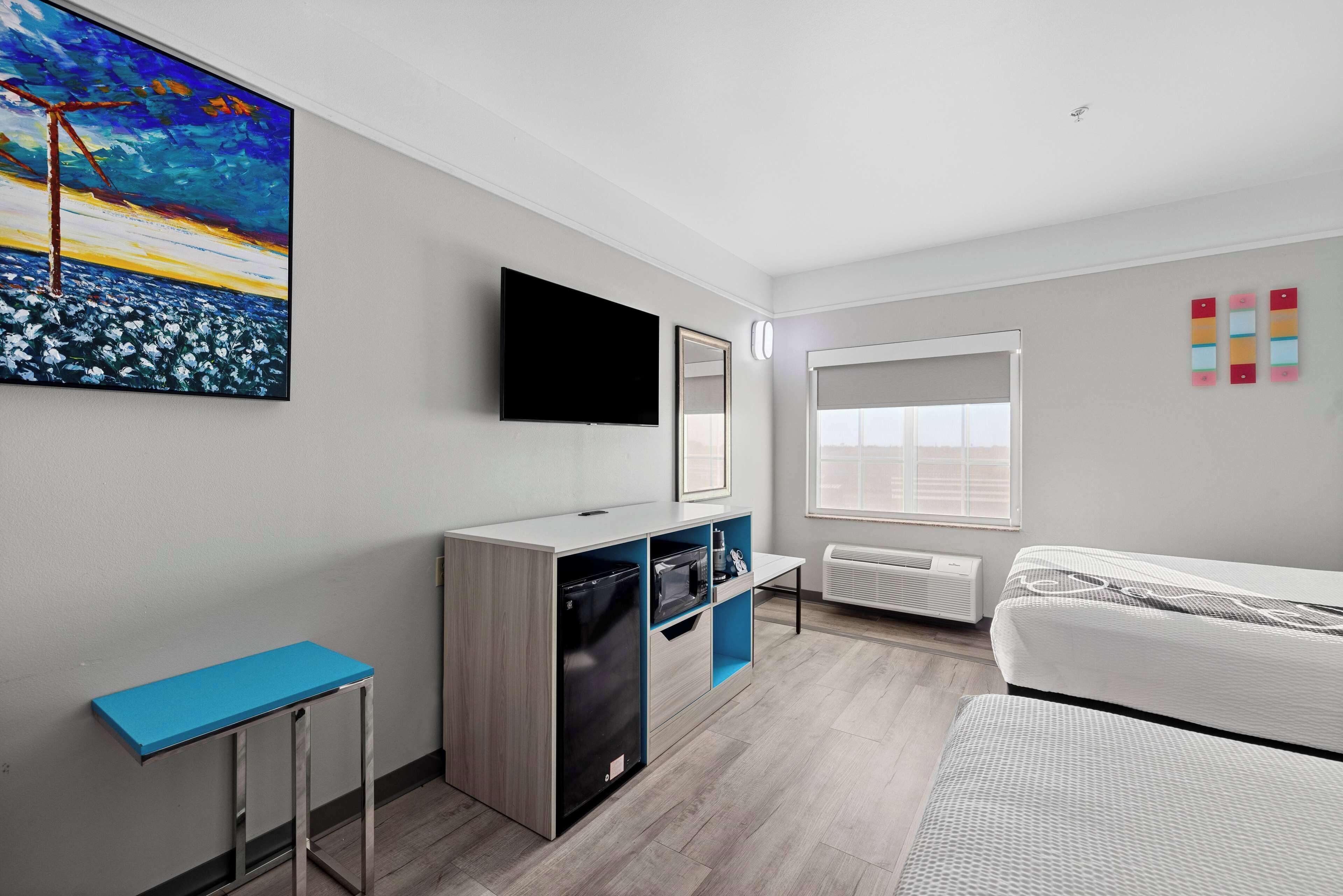 A modern hotel room with a bed, a desk, a flat-screen TV, a painting, and a window with blinds.