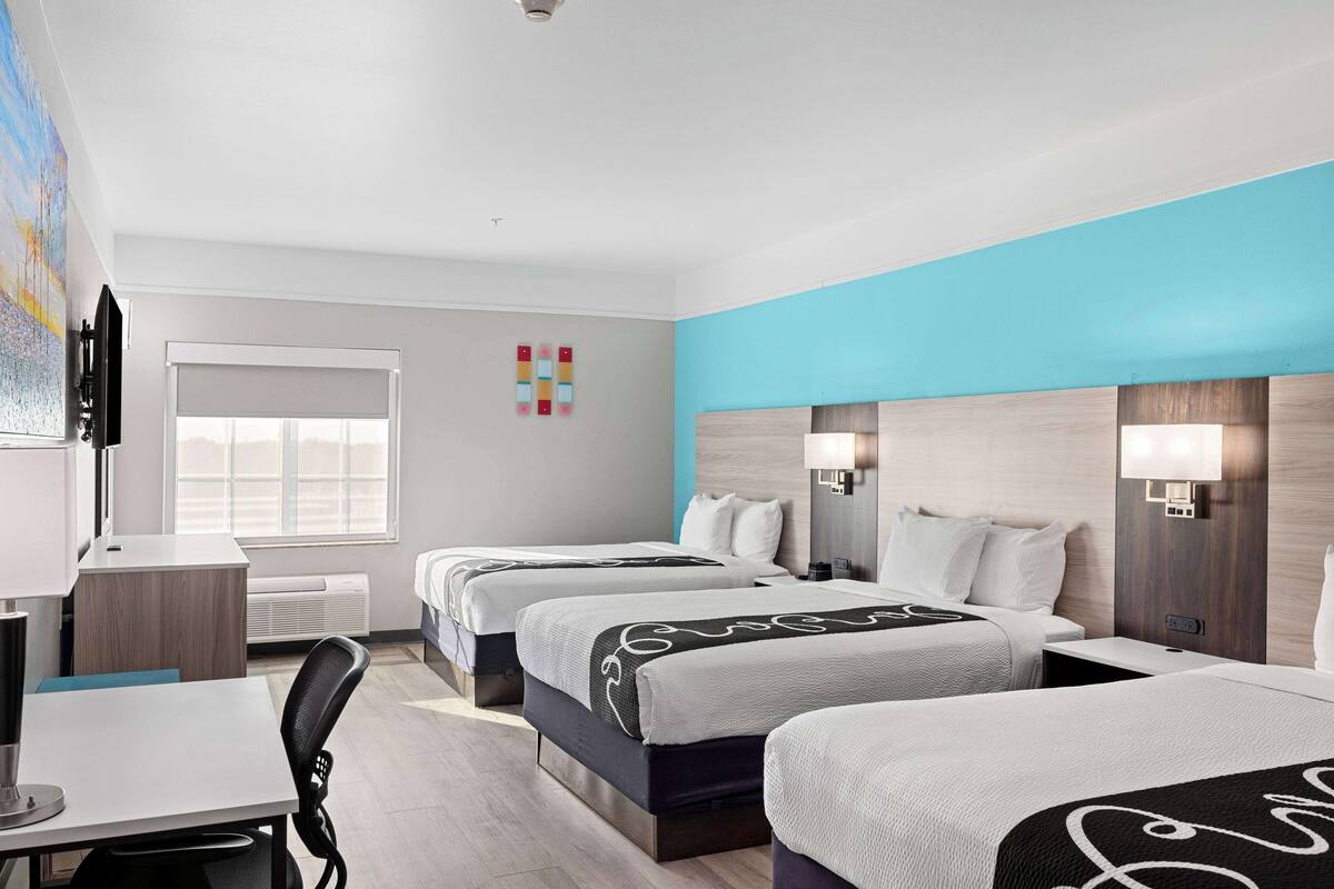 A hotel room with three beds, a desk, a chair, a TV, and a window with blinds.