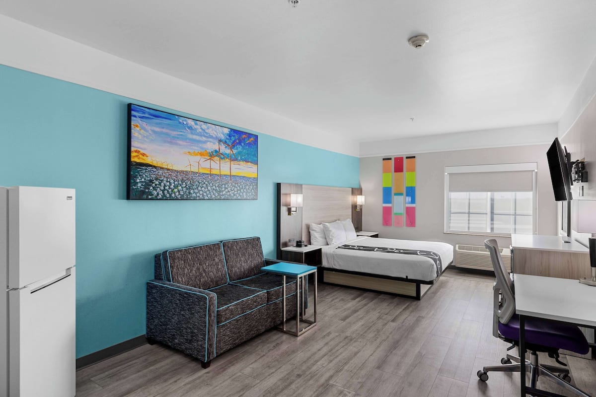 A modern hotel room with a blue accent wall, a painting, a bed, a desk, and a couch.