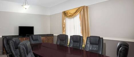 A conference room with a dark wooden table, black leather chairs, a flat-screen TV, and a large window with beige curtains.