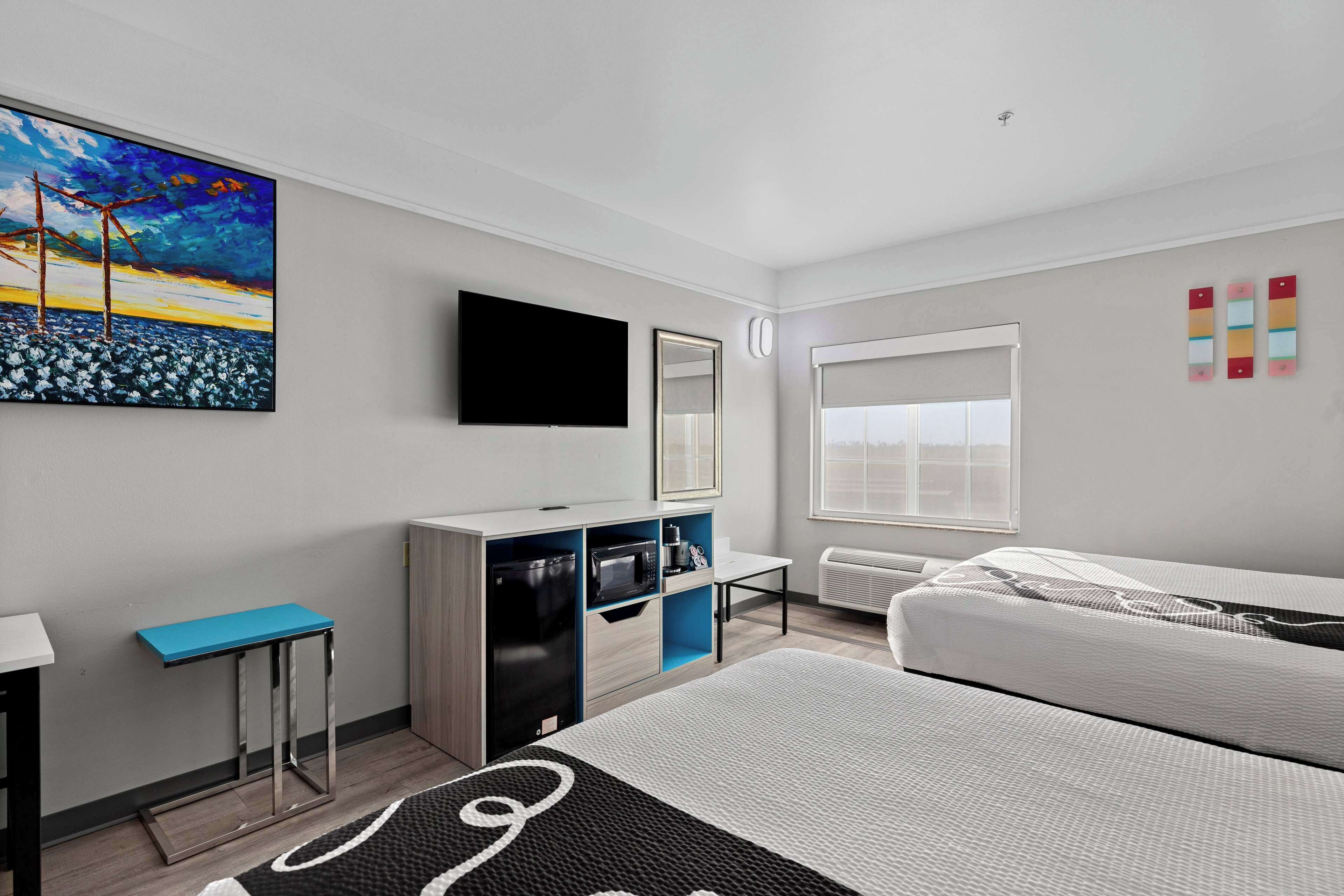 A modern hotel room with a bed, a desk, a flat-screen TV, a painting, and a window with blinds.