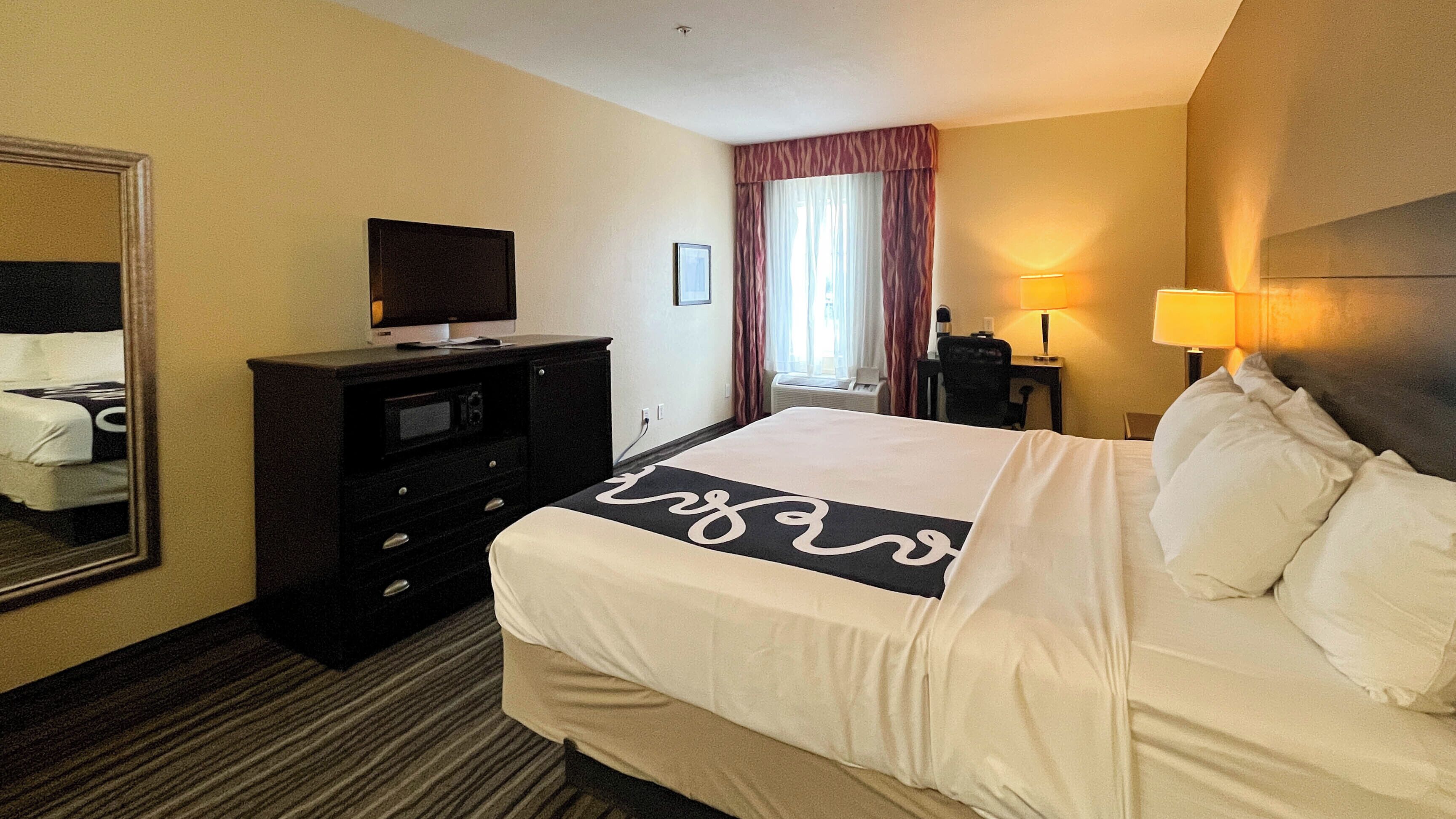 A hotel room with a bed, a television on a dresser, a desk with a lamp, and a window with curtains.