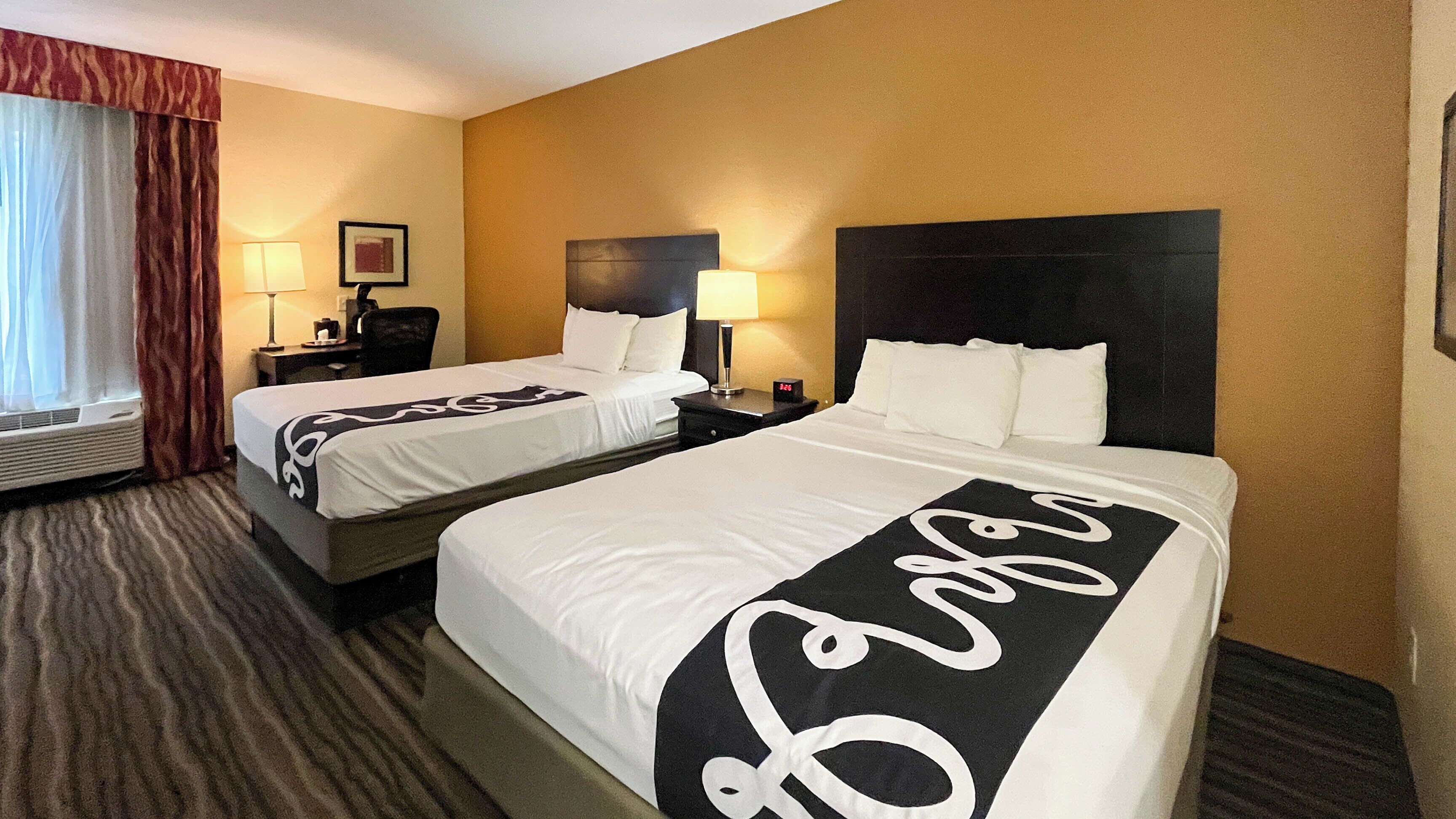 Habitación de hotel con dos camas, una mesita de noche con lámpara, un reloj y una ventana con cortinas.