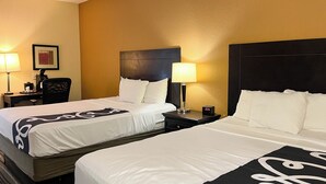 A hotel room with two beds, a nightstand with a clock, a desk with a lamp, and a chair.