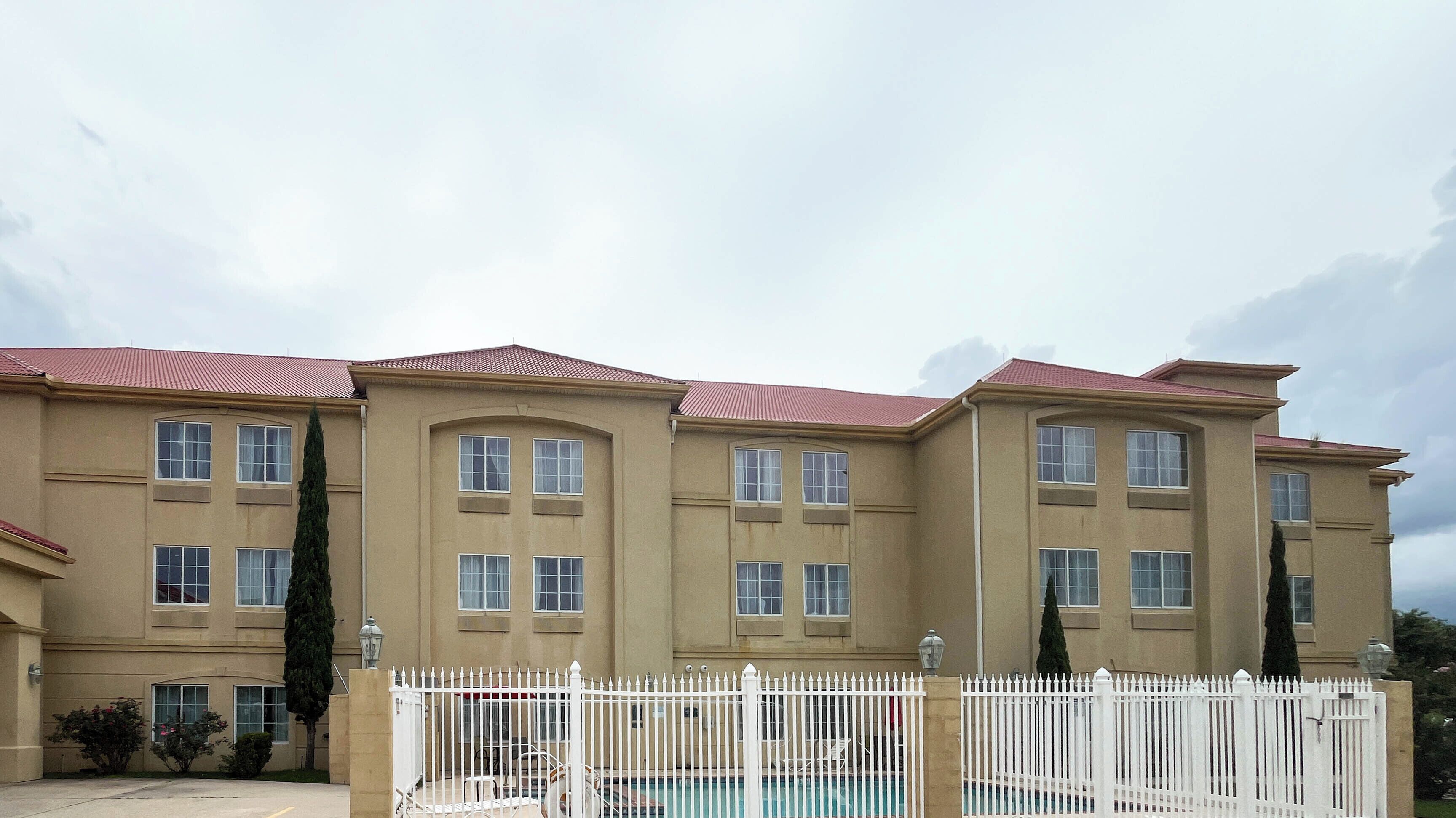 Un edificio a più piani con facciata beige, tetto in tegole rosse e un recinto bianco che circonda una piscina.